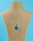 A sterling silver chain necklace with a round fused glass pendant featuring green, teal, and white wavy stripes, handmade in coastal colours and seen on a mannequin.