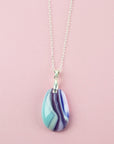 A teardrop-shaped fused glass pendant necklace with a marbled pattern of purple, blue, and light teal, suspended on a silver chain against a solid pink background.