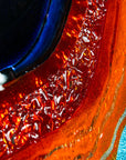 A detailed image of a circular fused glass wall panel featuring abstract wave design in ocean blues and bright oranges, with textured detailing.