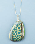 A handmade fused glass pendant necklace featuring a teardrop-shaped glass charm with a pattern of teal blue dots, bordered by brown and green swirls, suspended from a sterling silver chain.