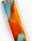 A close up of a fused glass hanging, hand-coloured in vibrant orange and bright aqua tones, with an orange ribbon.
