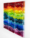 A three-panel vertical fused glass wall art piece featuring wavy horizontal rainbow stripes in red, orange, yellow, green, blue, and purple, mounted on a white wall.