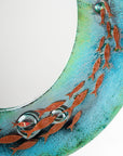 A close up of a circular fused glass round mirror, handmade in vibrant aquas and sea greens, with darting copper fish.