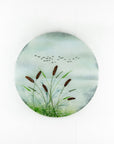 A circular fused glass wall art piece featuring a painted landscape with green reeds, brown seed heads, a pale blue sky, and small black birds, shown against a plain white background.