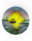 A fused glass circular wall art panel featuring a hand-painted design of a fisherman in a small boat casting a line in front of a large glowing yellow sun over a calm lake in natural, organic colours.