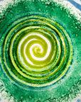 Close up of a fused glass round dish, featuring a dark emerald colour-way and a textured swirling detailing.