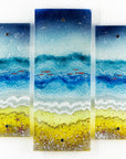 Fused glass triptych depicting a beach scene with blue water, yellow sand, and red, copper fish.