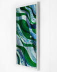 Side on image of a fused glass oblong art frame with coastal greens and textural blues. Hand-made with lass fish layered on and a white frame.