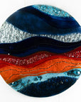 A circular fused glass wall panel featuring abstract wave design in ocean blues and bright oranges, with textured detailing.