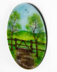 A side view circular fused glass wall art piece featuring a painted landscape with green trees, a wooden fence, rolling hills, and a dirt path under a light blue sky.