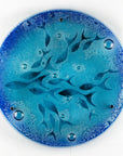 A round fused glass wall panel featuring hand-cut glass fish and bubbles, designed in a marine blue colour-way.