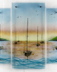 A fused glass triptych depicting a serene harbour scene with hand-painted sailboats on blue water under a pastel sky with hills in the distance.