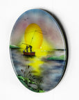 A fused glass circular wall art panel featuring a hand-painted design of a fisherman in a small boat casting a line in front of a large glowing yellow sun over a calm lake in natural, organic colours.