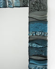 A close up of a fused glass oblong mirror, handmade using soft coastal blue tinted and grey glass with delicate texture and a wavy design.