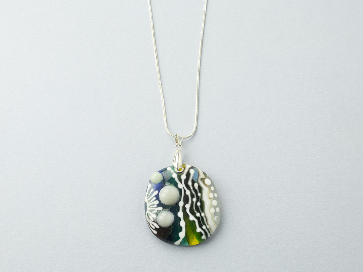 A fused glass pendant necklace, handmade in soft coastal and organic hues, with hand-painted botanical patterns, hung on sterling silver chain. 