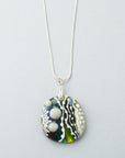 A fused glass pendant necklace, handmade in soft coastal and organic hues, with hand-painted botanical patterns, hung on sterling silver chain. 