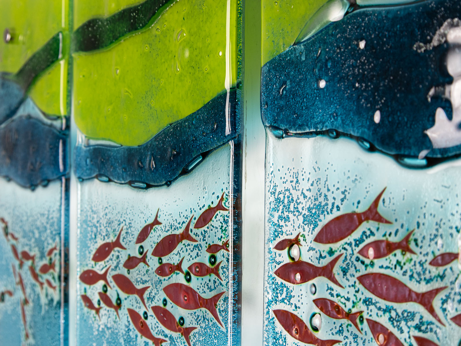 A close up of a fused glass triptych featuring marine blues and greens in an ocean design, with copper fish and textured detail. 