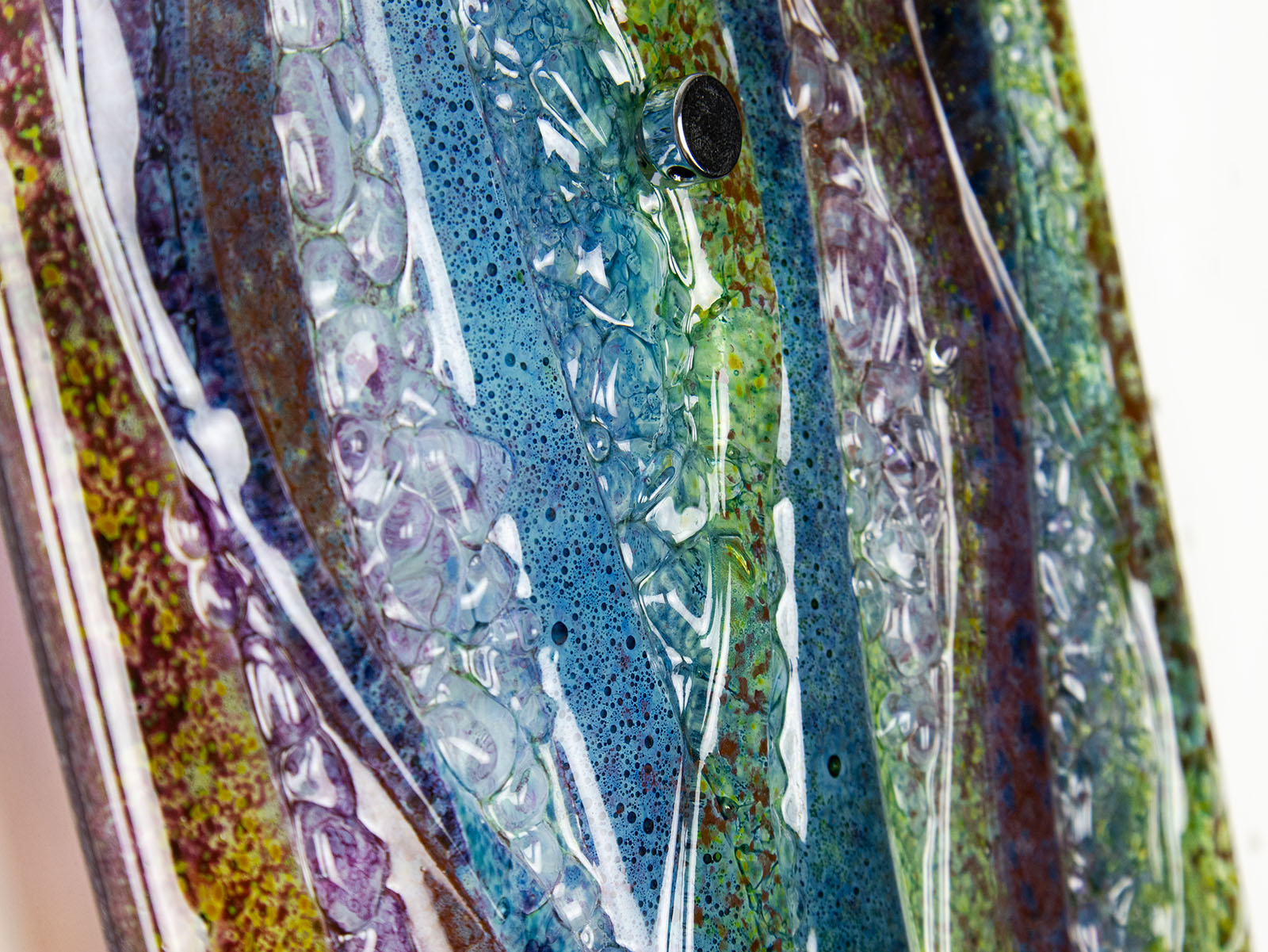 A close up or organic texture on a fused glass wall panel, featuring a wavy design in sea blues, greens and amethyst purples with textured detailing. 