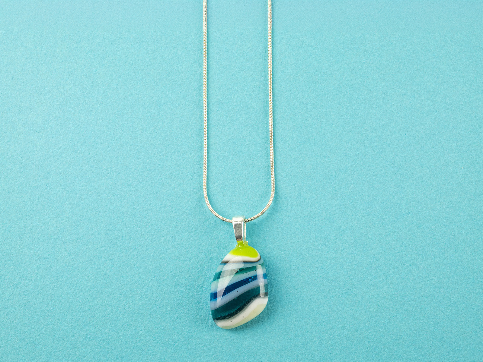 A teardrop-shaped fused glass pendant necklace with a marbled pattern of green, blue, and light teal, suspended on a sterling silver chain.
