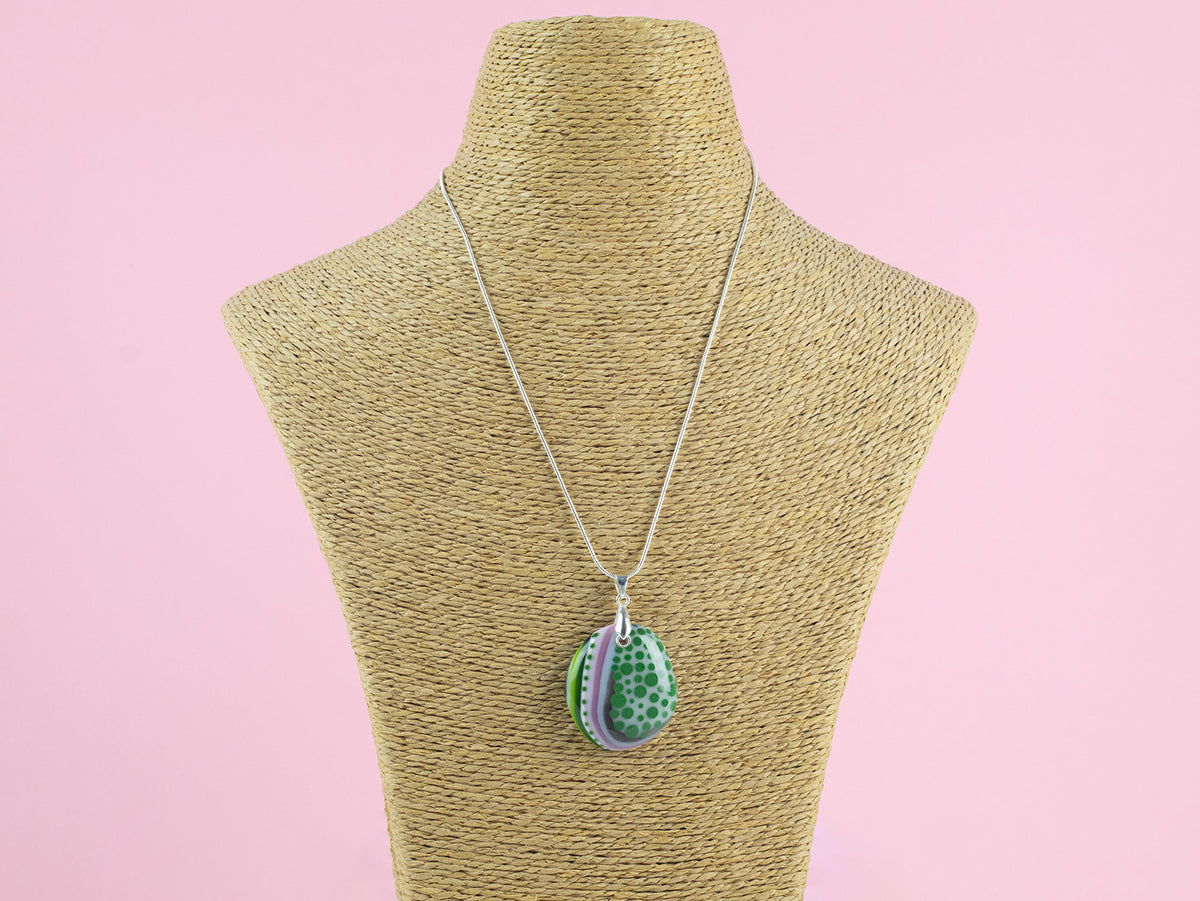 A fused glass pendant necklace, handmade in sea greens and blushed pinks, with hand-painted patterns, hung on a sterling silver chain. 