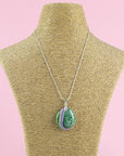 A fused glass pendant necklace, handmade in sea greens and blushed pinks, with hand-painted patterns, hung on a sterling silver chain. 