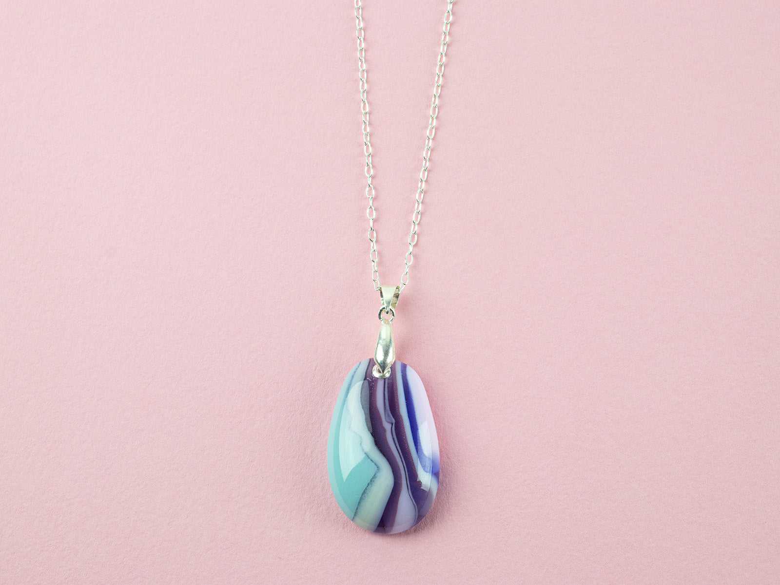 A teardrop-shaped fused glass pendant necklace with a marbled pattern of purple, blue, and light teal, suspended on a silver chain against a solid pink background.