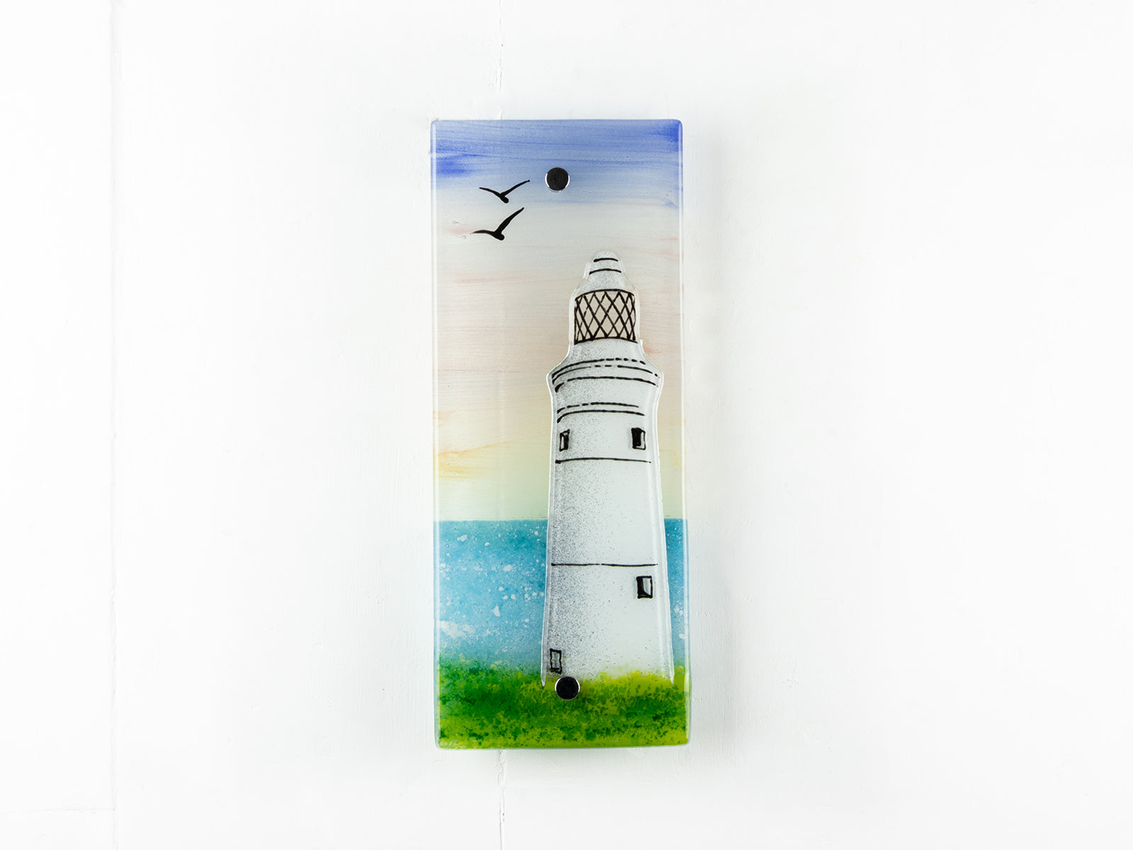A fused glass extra small wall panel, featuring a lighthouse design with a very soft sunset pink sky, with soaring birds. A soft coastal piece of wall art. 