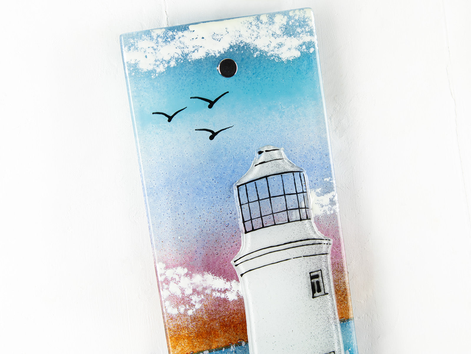 A close up of a fused glass extra small wall panel, featuring a lighthouse design with a bright sunset pink sky, with soaring birds. A soft coastal piece of wall art. 