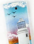 A close up of a fused glass extra small wall panel, featuring a lighthouse design with a bright sunset pink sky, with soaring birds. A soft coastal piece of wall art. 