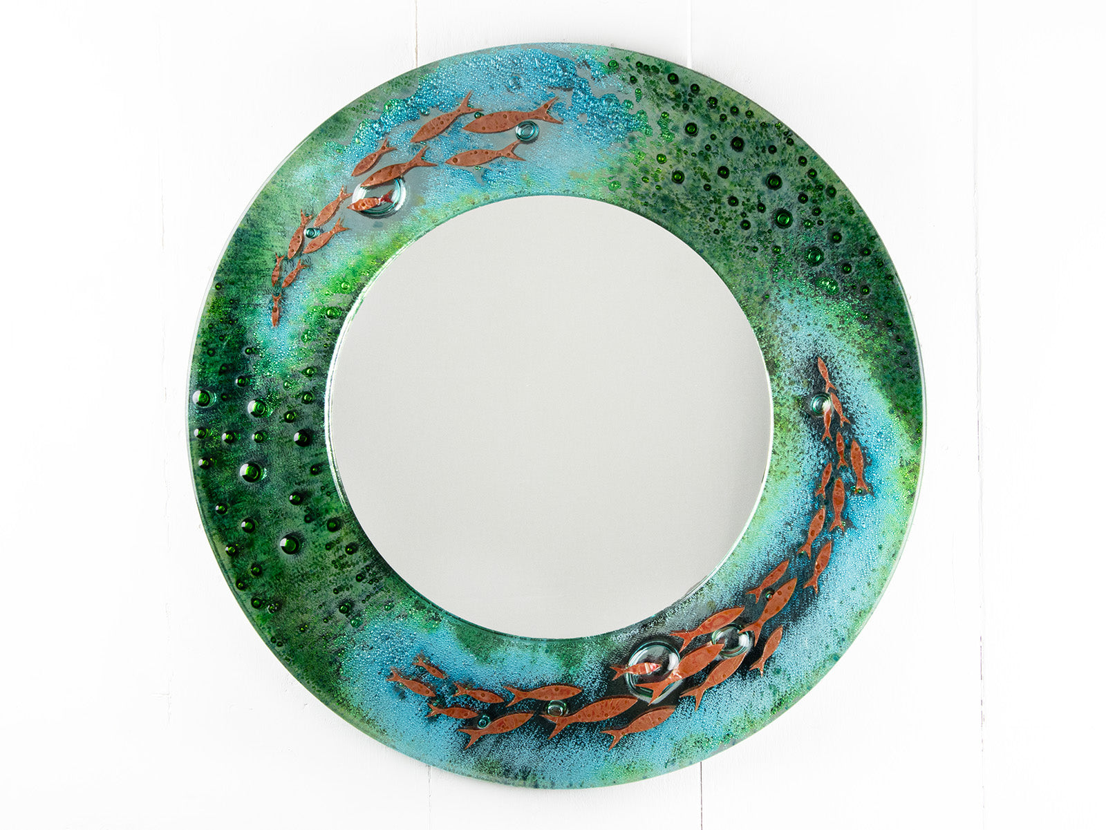 A circular fused glass round mirror, handmade in vibrant aquas and sea greens, with darting copper fish. 