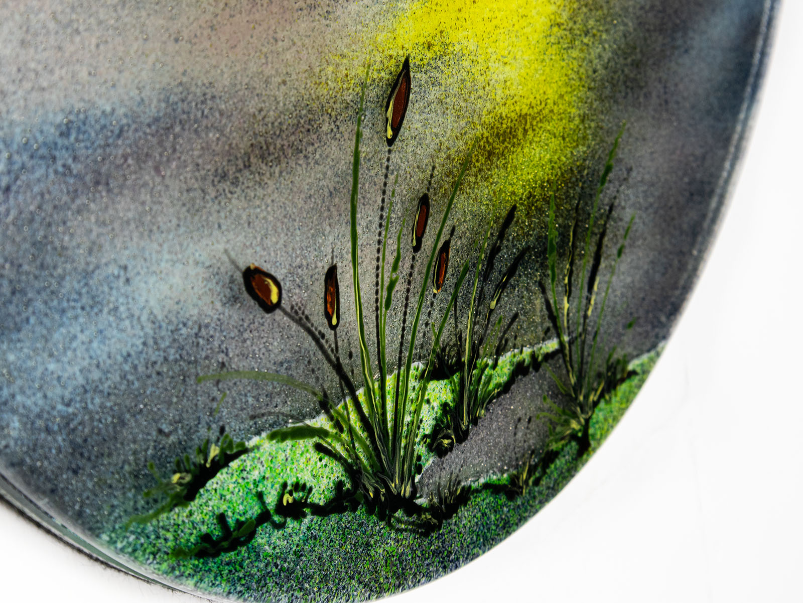 A close up of the grass detailing on a fused glass circular wall art panel featuring a hand-painted design of a fisherman in a small boat casting a line in front of a large glowing yellow sun over a calm lake in natural, organic colours. 