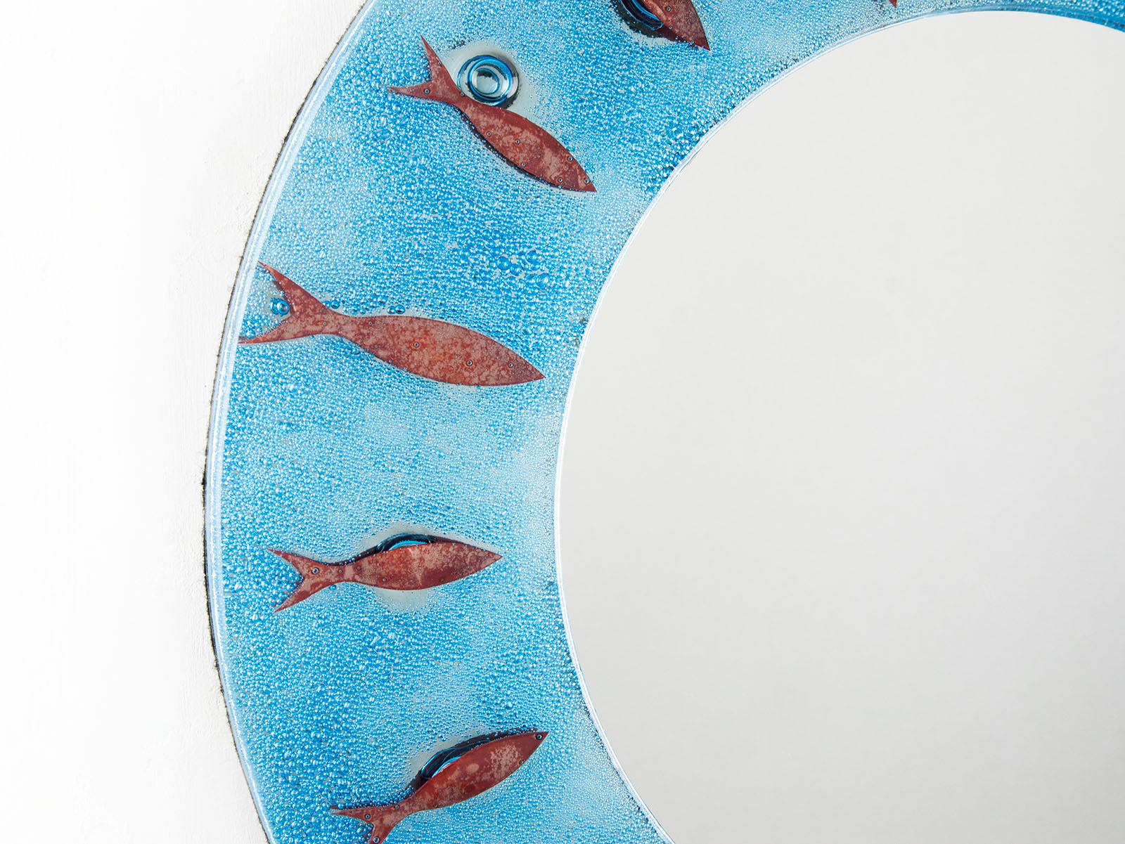 A close up of a fused glass round mirror in an aqua colour-way featuring a circular design of copper fish.