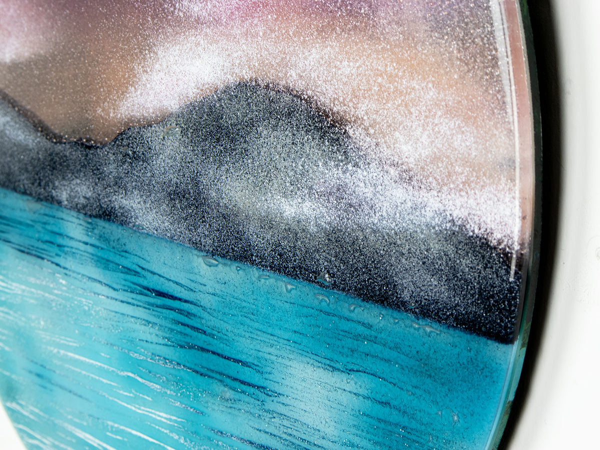 A close up of a circular wall art fused glass panel featuring a scenic landscape with a blue and purple sky, misty mountains, and turquoise water. 