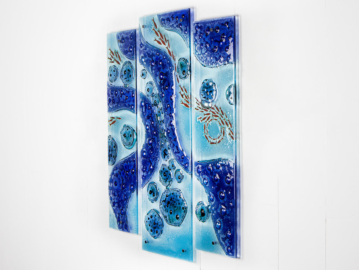 A fused glass staggered triptych wall display, handmade in bright ocean blues and aquas, in a coastal rock-pool design, featuring copper fish. 
