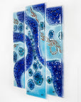 A fused glass staggered triptych wall display, handmade in bright ocean blues and aquas, in a coastal rock-pool design, featuring copper fish. 