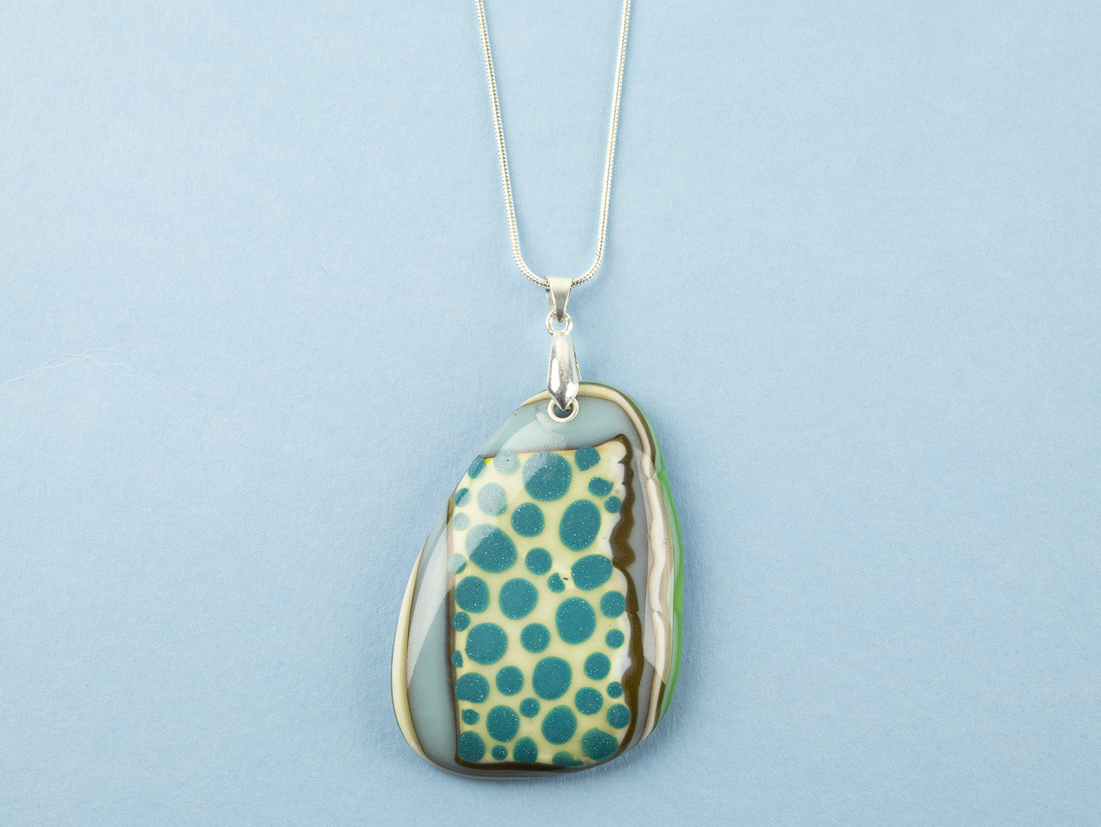 A handmade fused glass pendant necklace featuring a teardrop-shaped glass charm with a pattern of teal blue dots, bordered by brown and green swirls, suspended from a sterling silver chain.