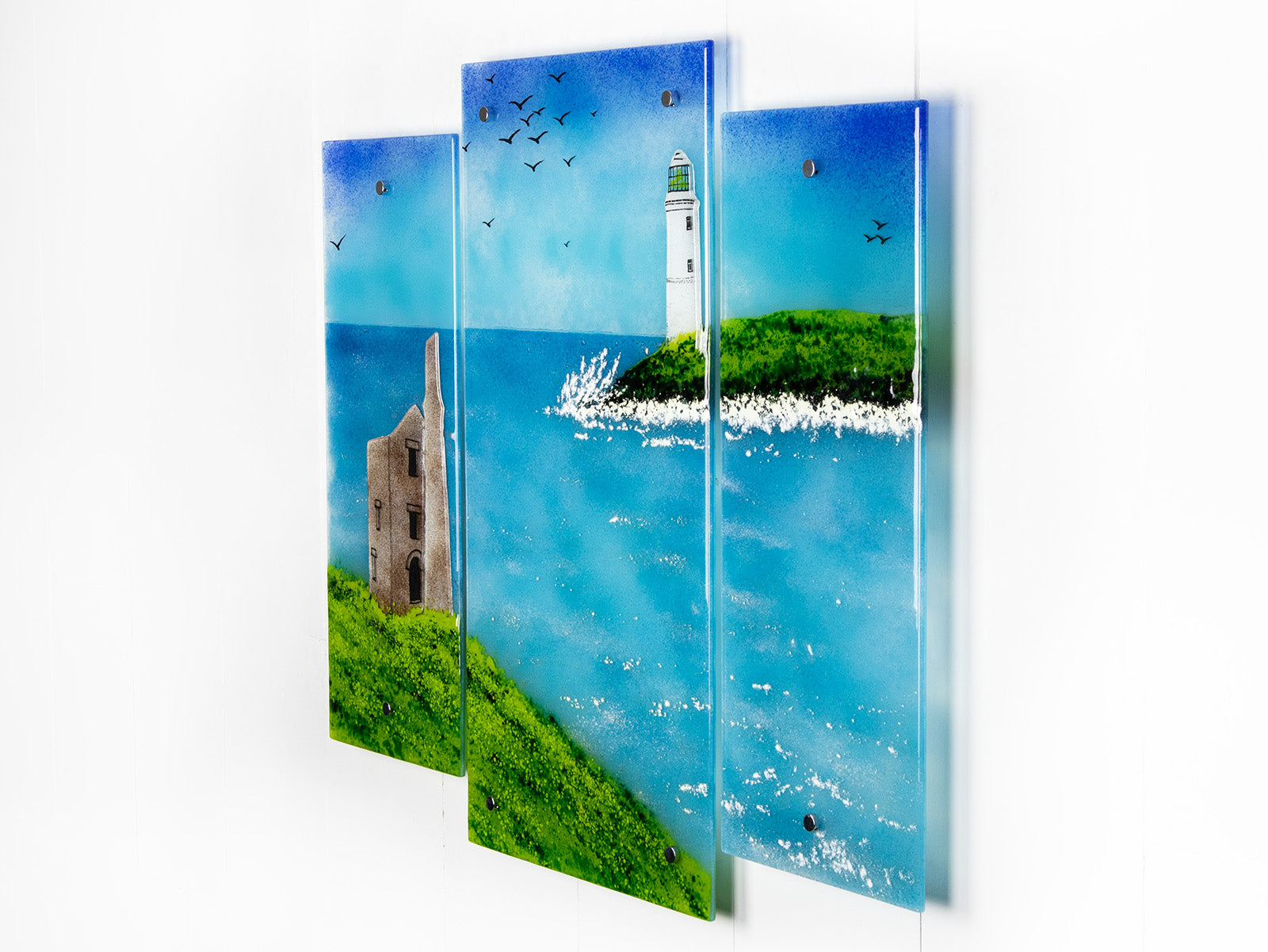 A fused glass triptych in coastal colours, with distant soaring sea birds, a lighthouse sat on a cliff and a Cornish tin mine. Hand-made coastal wall art, with texture. 