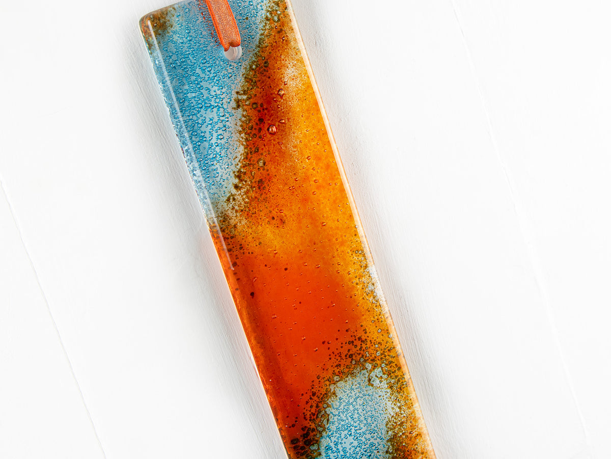 A close up of a fused glass hanging, hand-coloured in vibrant orange and bright aqua tones, with an orange ribbon. 