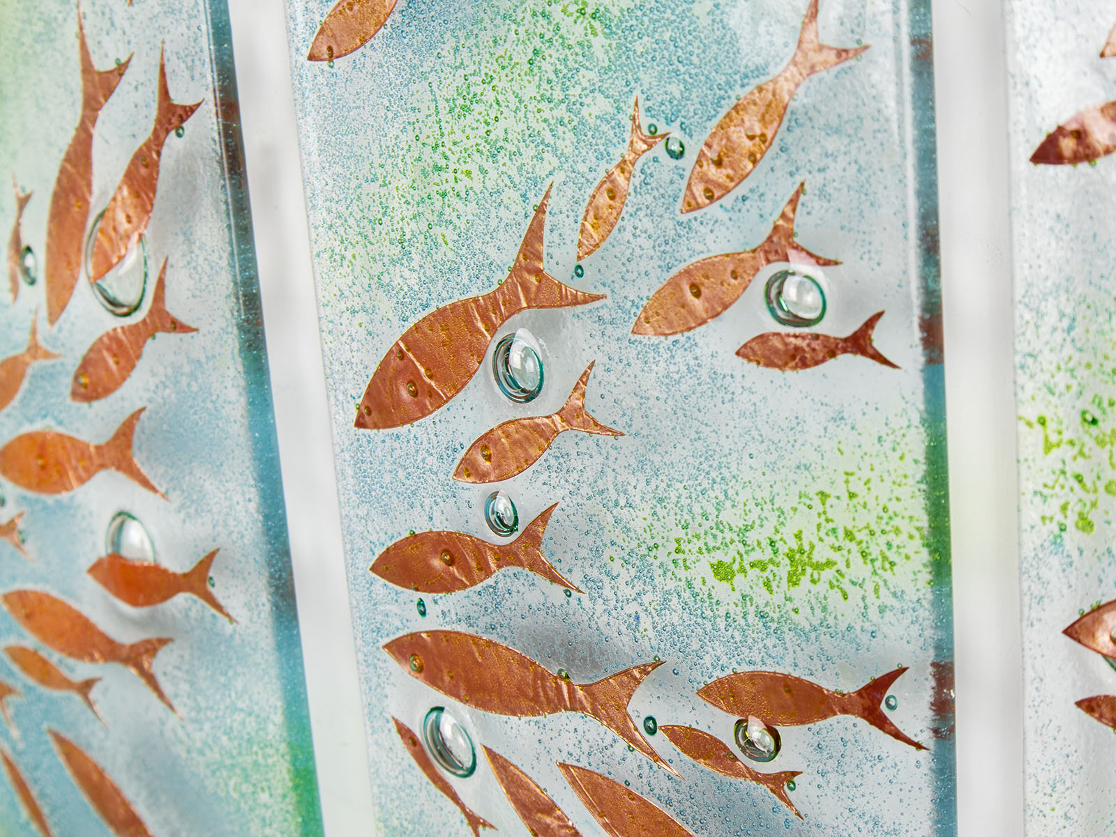 A close-up of a fused glass quintych featuring a soft coastal colour-way with darting copper fish and bubbled detailing. An ocean inspired piece of wall art. 