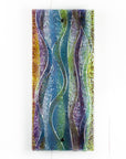 A fused glass wall panel, featuring a wavy design in sea blues, greens and amethyst purples with textured detailing. 