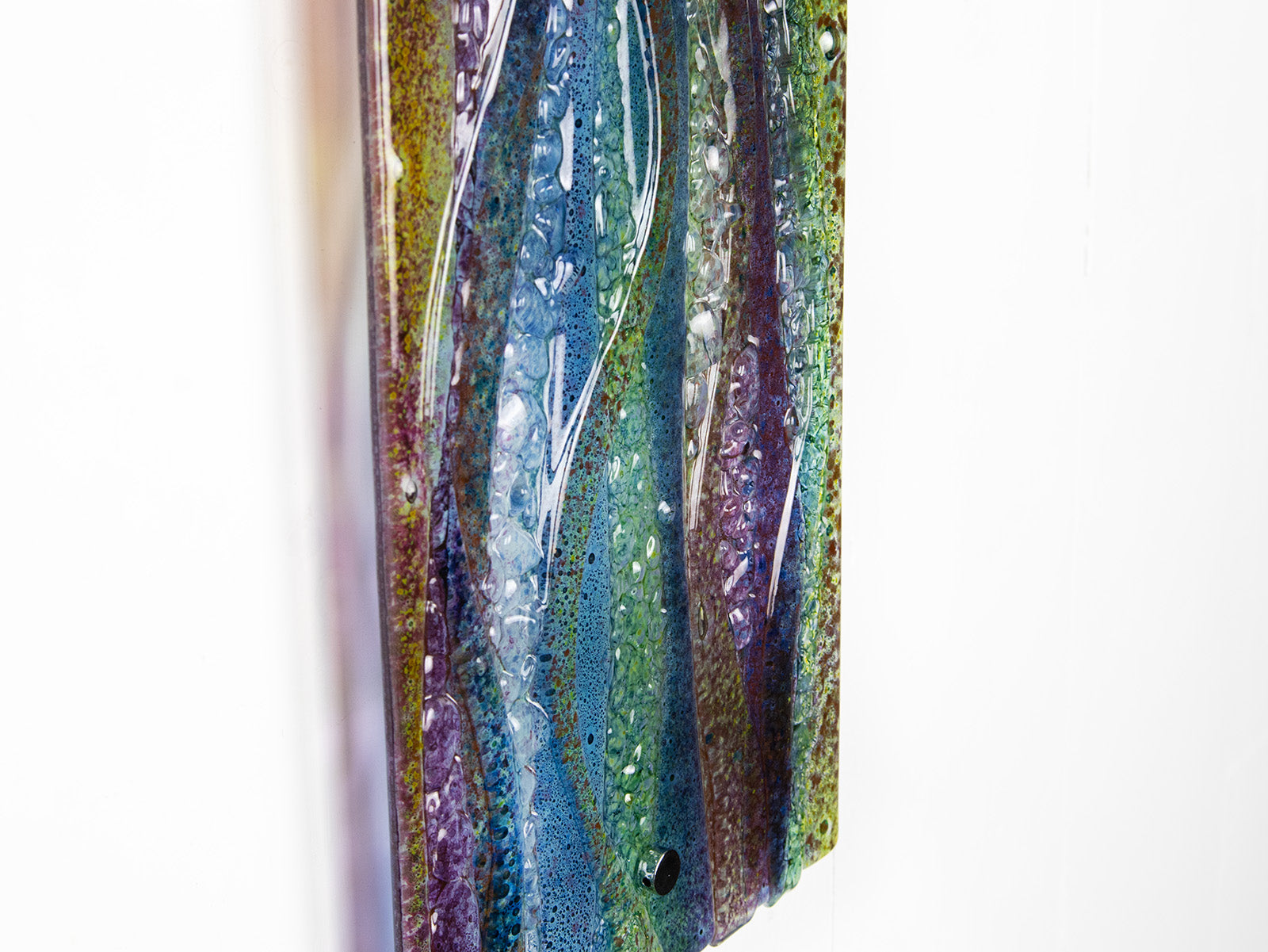 A close up of a fused glass wall panel, featuring a wavy design in sea blues, greens and amethyst purples with textured detailing. 