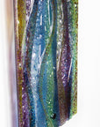 A close up of a fused glass wall panel, featuring a wavy design in sea blues, greens and amethyst purples with textured detailing. 