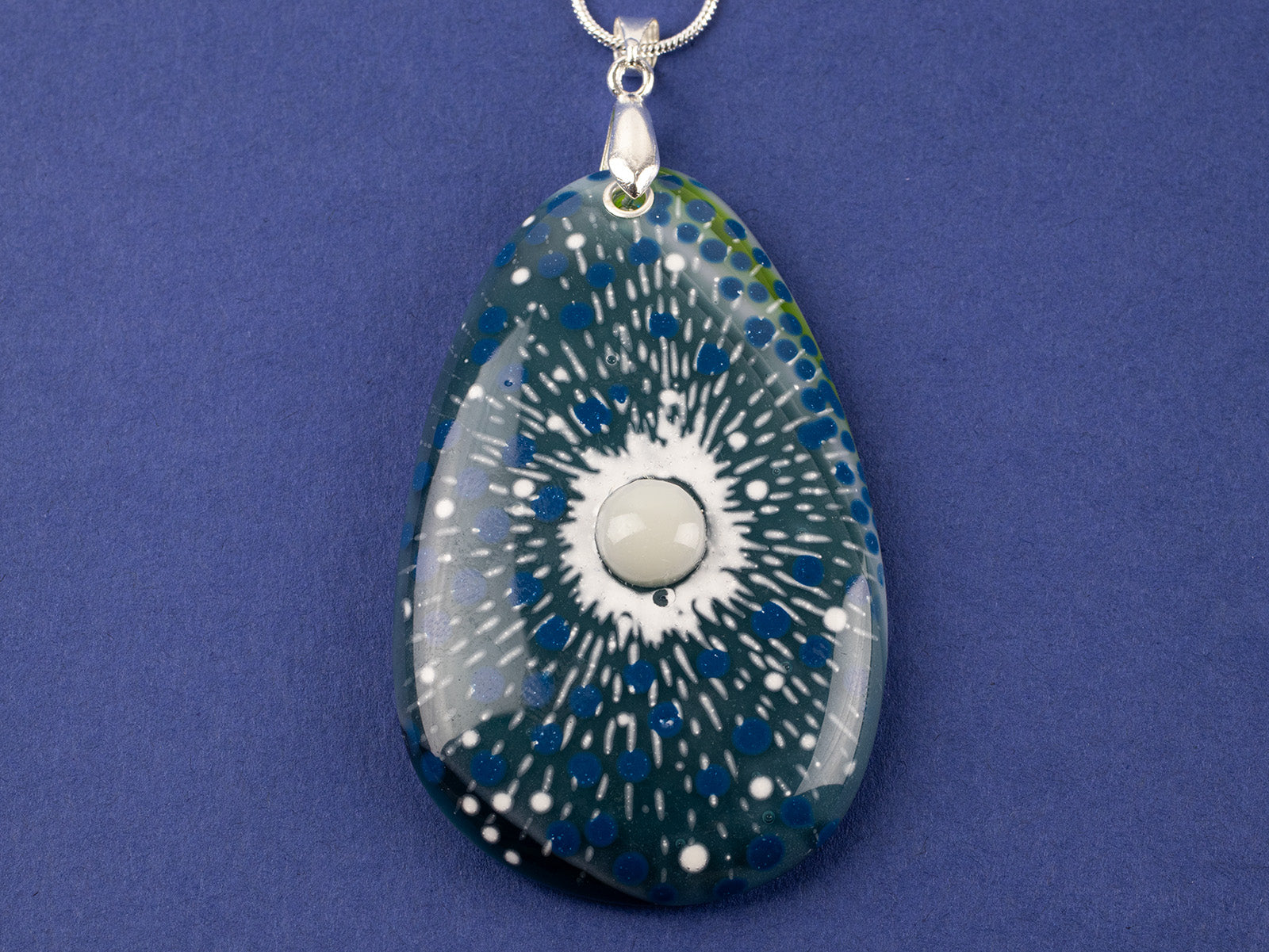 A close up of a teardrop-shaped fused glass pendant necklace featuring a blue and green sunburst pattern with white speckles and a central pearl inspired disc, suspended from a sterling silver chain.