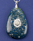 A close up of a teardrop-shaped fused glass pendant necklace featuring a blue and green sunburst pattern with white speckles and a central pearl inspired disc, suspended from a sterling silver chain.