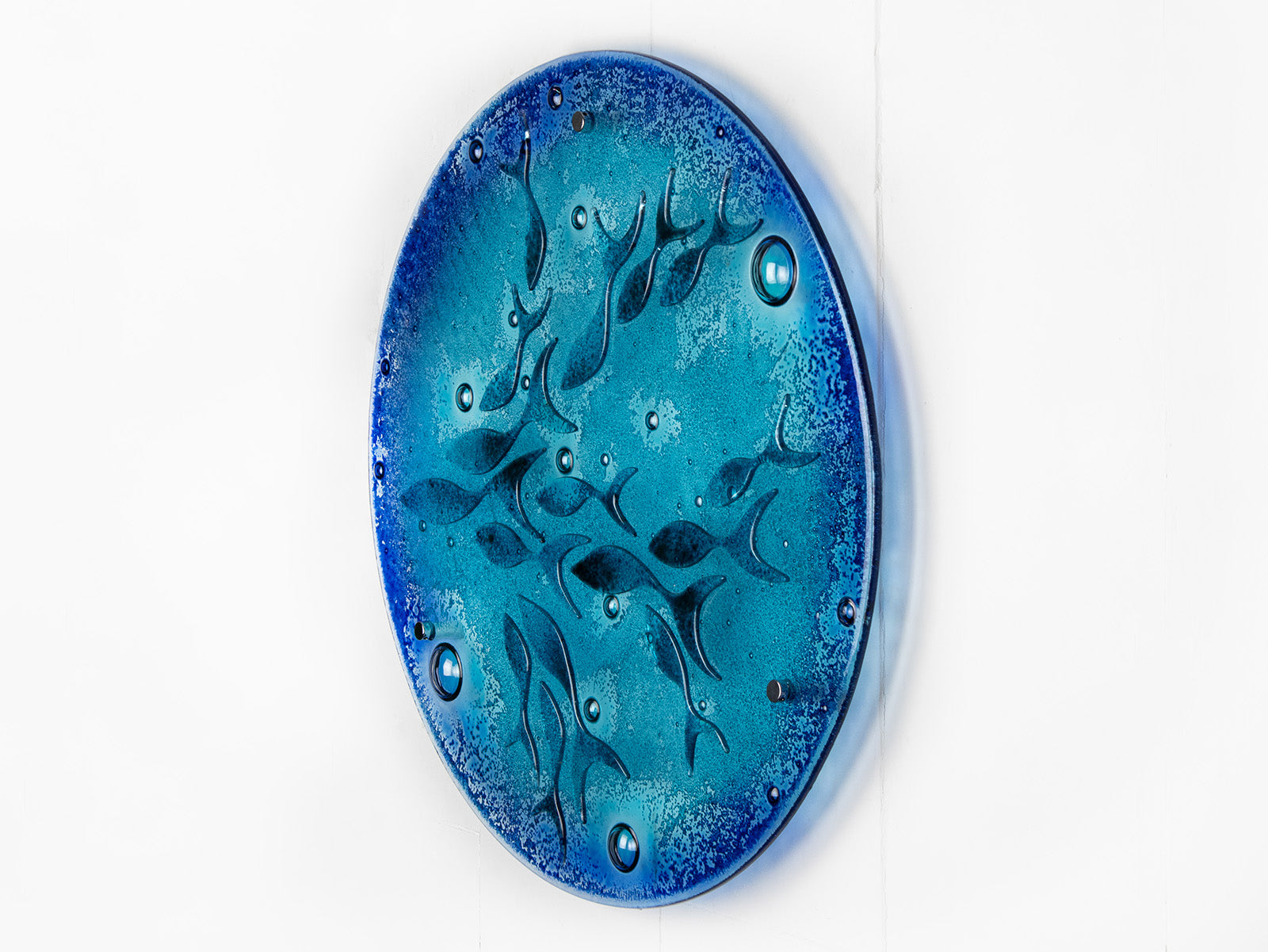 A side view of a round fused glass wall panel featuring hand-cut glass fish and bubbles, designed in a marine blue colour-way.