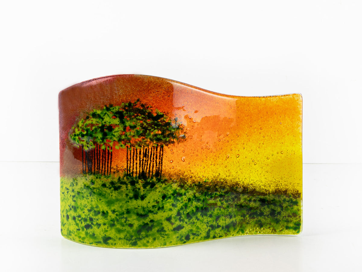 A fused glass wave sculpture, with a countryside design on a cluster of trees on a grassy hill, with a sunset sky behind. 