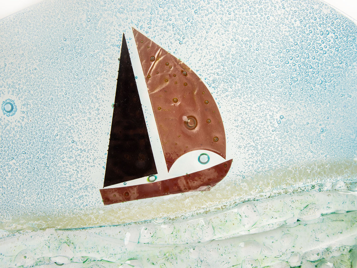 A close up of a sail boat designed on a fused glass wave sculpture with a metal sail boat and a soft coastal colour-way, with textured details. 