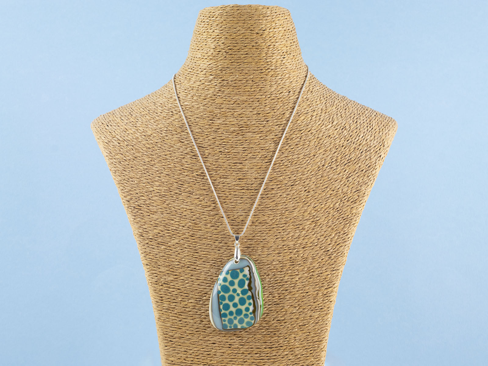 A handmade fused glass pendant necklace featuring a teardrop-shaped glass charm with a pattern of teal blue dots, bordered by brown and green swirls, suspended from a sterling silver chain.