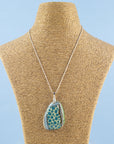 A handmade fused glass pendant necklace featuring a teardrop-shaped glass charm with a pattern of teal blue dots, bordered by brown and green swirls, suspended from a sterling silver chain.