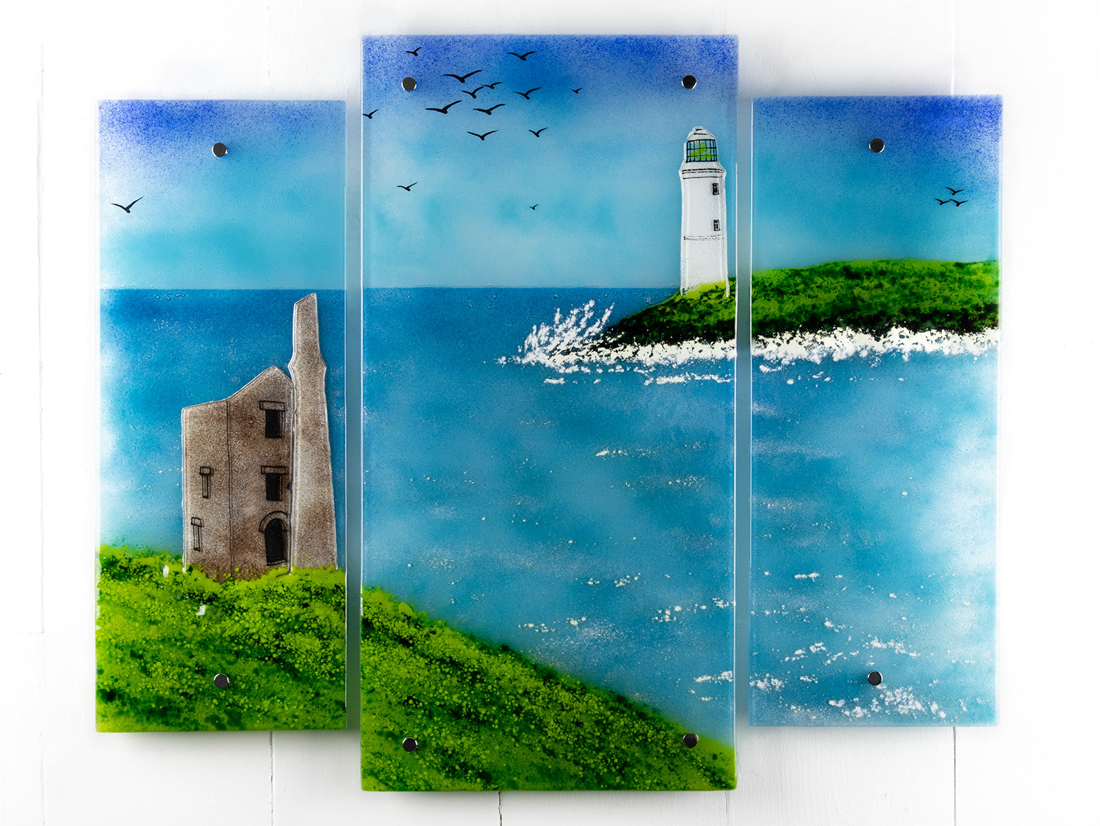 A fused glass triptych in coastal colours, with distant soaring sea birds, a lighthouse sat on a cliff and a Cornish tin mine. Hand-made coastal wall art, with texture. 